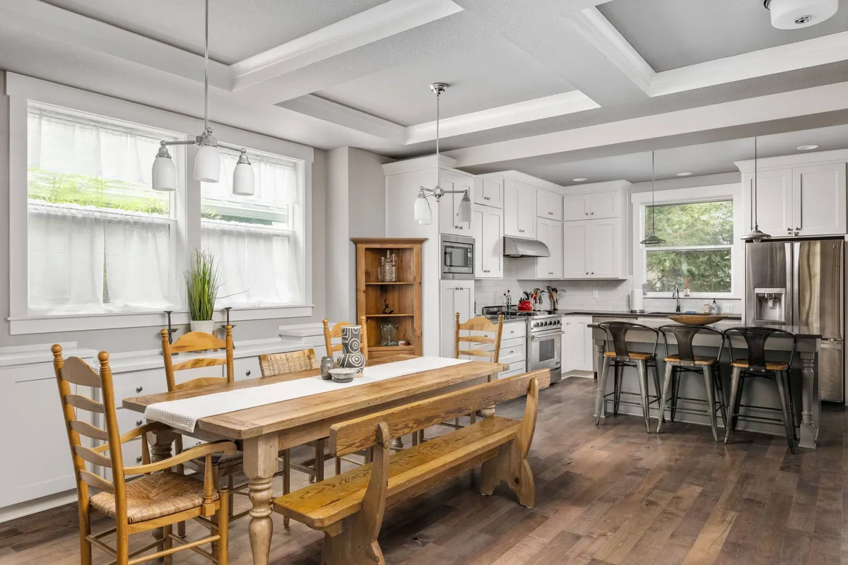 This image shows a large wooden dining table paired with a modern kitchen island, surrounded by windows draped with sheer curtains that allow sunlight to fill the space. Stainless steel appliances and well-crafted cabinetry provide a stylish, functional backdrop. The setting feels open and inviting, ideal for shared meals and lively gatherings.