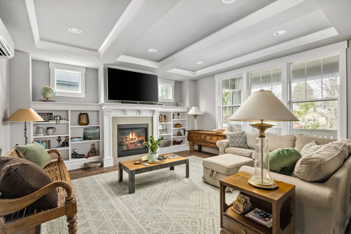 The image showcases a beautifully styled living room with a crackling fireplace as the centerpiece, plush sofa, a warm coffee table, and sunlight pouring through large windows. The ambiance is cozy and welcoming, perfect for relaxing after exploring Portland or gathering with loved ones.