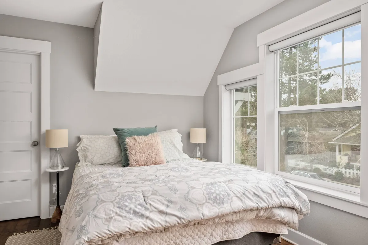 A bedroom with decorative pillows on the bed, large windows flooding the space with natural light, and a nightstand with a lamp. The bright, tranquil atmosphere suggests a peaceful return after a day spent outdoors, and the window emphasizes the connection to the outside neighborhood.