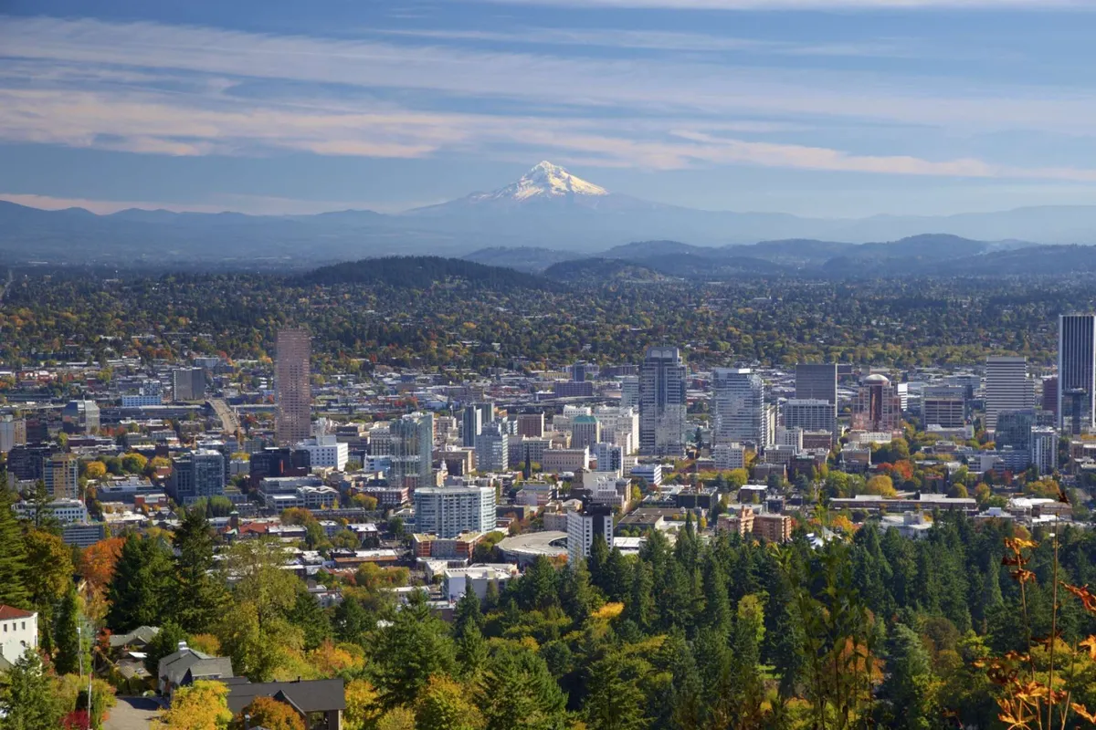 A panoramic view of downtown Portland with Mount Hood looming in the distance, framed by lush greenery. The city is bustling yet serene, inviting visitors to explore or simply enjoy the view.
