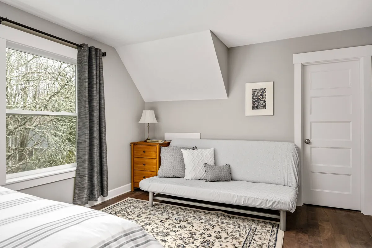 A cozy living space featuring a sofa bed, wooden dresser, lamp, and a window with curtains. The room is bathed in gentle light, creating a welcoming spot for socializing, relaxing, or lounging with a pet after a day out in the neighborhood.