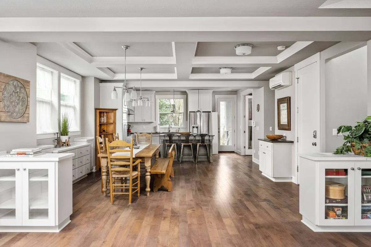The photo presents a warm, sunlit interior featuring a dining table adjacent to a kitchen island, all framed by large windows that bathe the room in natural light. The kitchen is modern and functional, inviting family and friends to cook together or enjoy conversation over a meal.