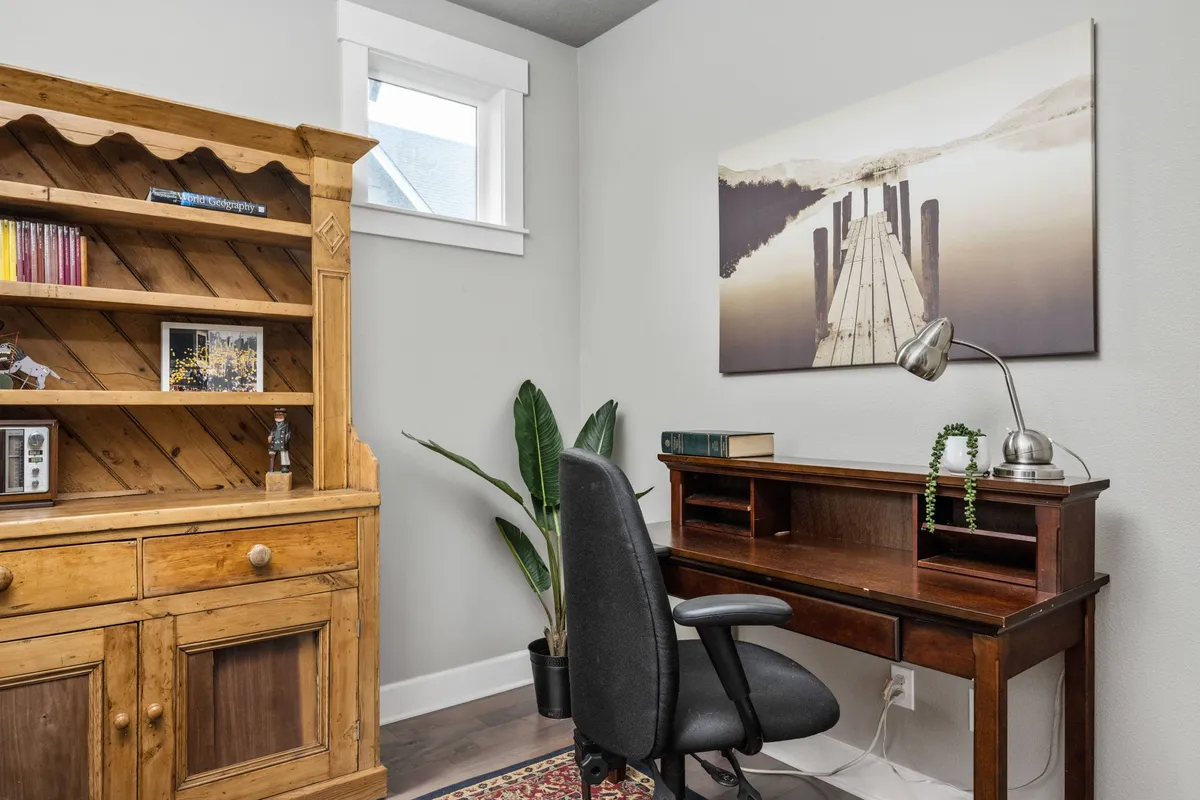 A serene workspace featuring a wooden bookshelf filled with books, a desk with a comfortable chair positioned for focus, and calming wall art depicting a dock. The area is bathed in natural light, creating a peaceful and productive environment ideal for work or study.