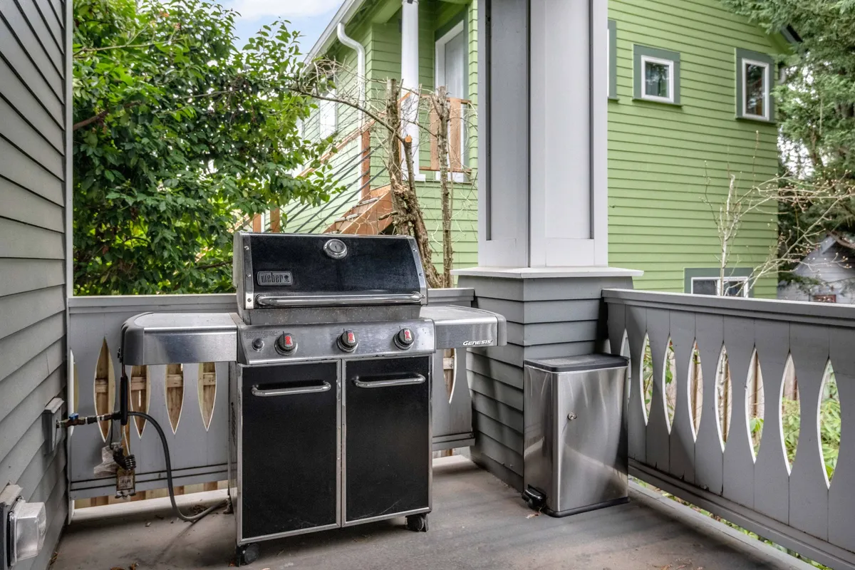 A vibrant backyard patio setting highlighted by a modern grill, sturdy railing, and the side of a green house. The space is clearly set up for outdoor entertaining, with the grill as a focal point for barbecues and social gatherings, and offers a relaxed atmosphere for spending time outdoors.