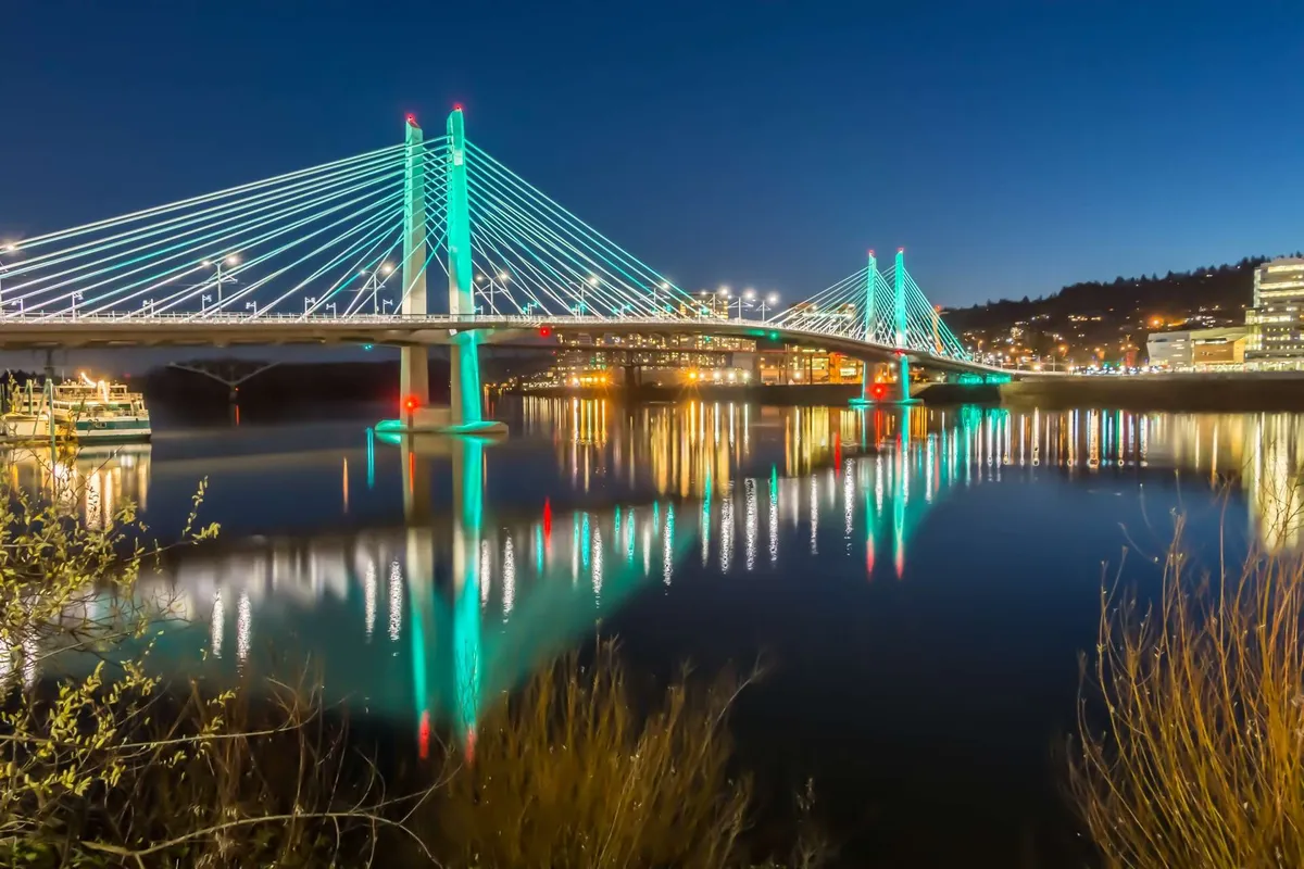 A scenic evening exterior shot featuring a suspension bridge illuminated by city lights, with vibrant reflections dancing on the river below. The surrounding cityscape hints at lively urban life, with restaurants and nightlife nearby.