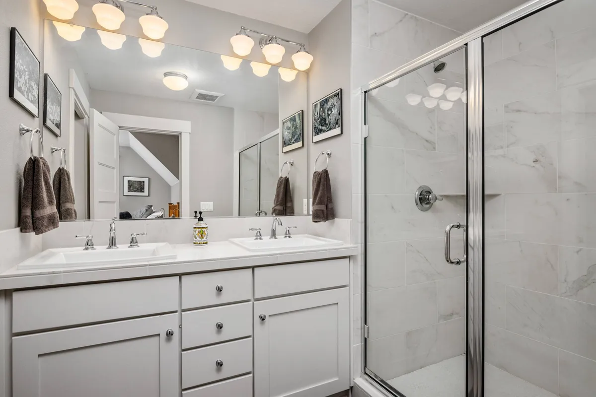 An airy, modern bathroom featuring a double sink vanity, a shower enclosure, a well-lit mirror, and tasteful framed artwork. The space feels fresh and inviting—designed for both daily routines and moments of self-care.