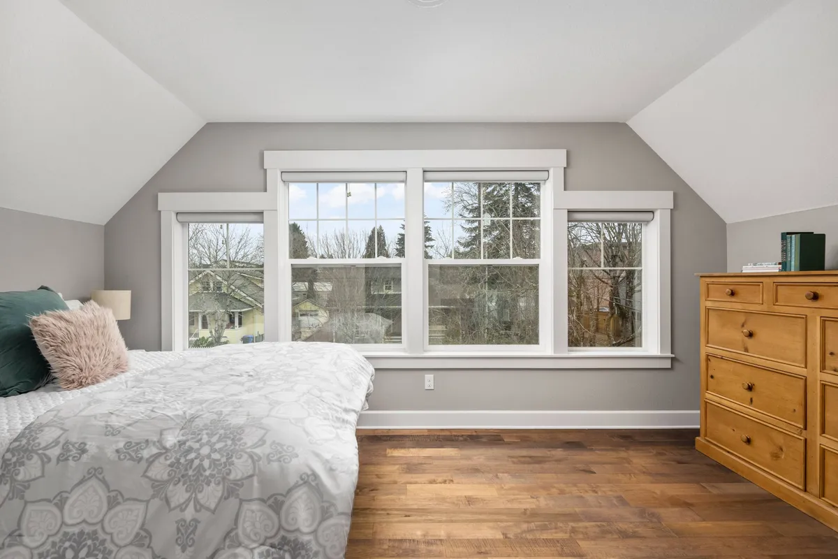 A serene queen bedroom outfitted with a comfortable bed adorned with decorative throw pillows. Large windows offer abundant natural light and peaceful views, while a wooden dresser adds a practical touch. The atmosphere is calm and restful, ideal for unwinding after a long day.